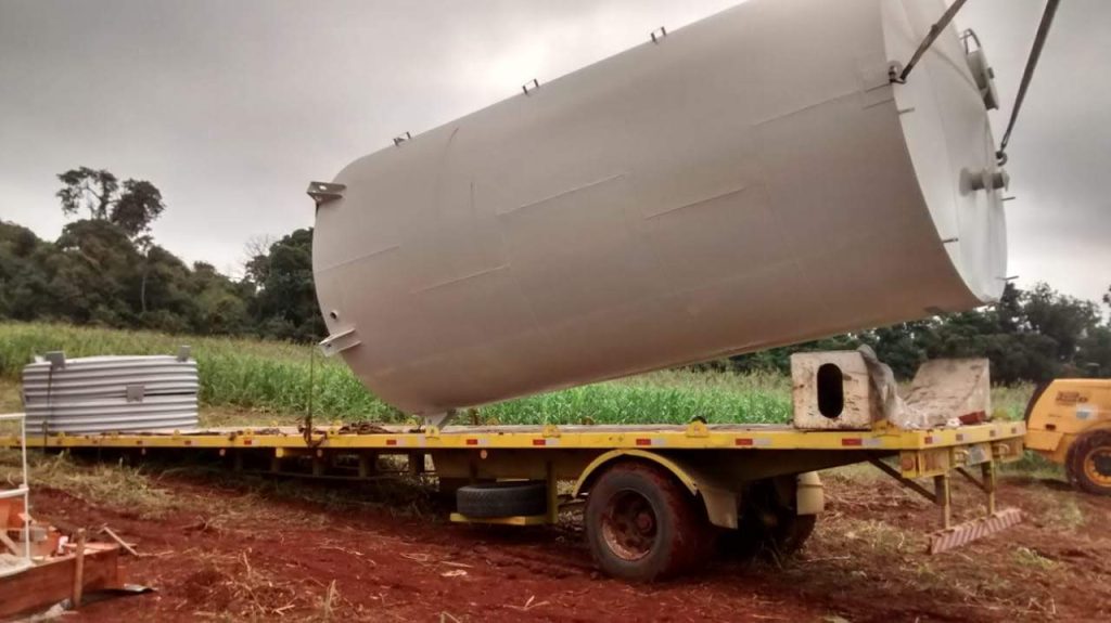 Transporte de grande caixa d'água para fazenda em caminhão prancha, mostrando o reservatório metálico sendo posicionado em solo de terra.