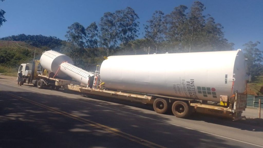 Caminhão transportando reservatório FNDE de grande porte em rodovia, utilizado para armazenamento de água em projetos públicos.