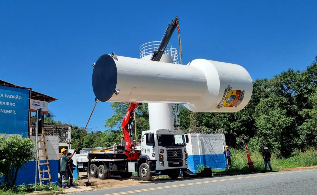 Caminhão munck realizando o transporte e manuseio de um reservatório tipo taça metálico branco em uma área urbana com vegetação.